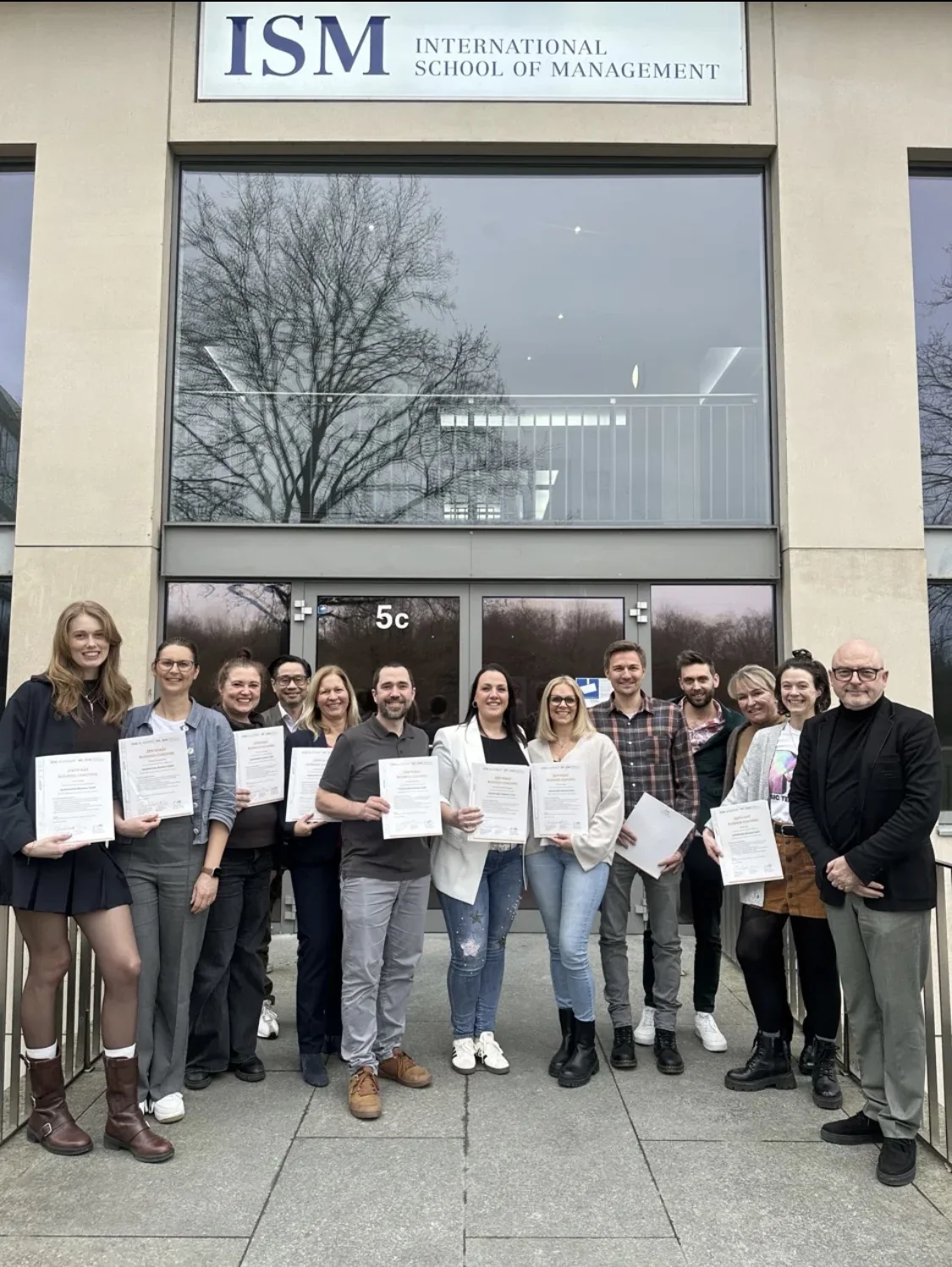 Absolventinnen und Absolventen der Business Coach Ausbildung mit ihren Zertifikaten vor dem Eingang der International School of Management in Köln