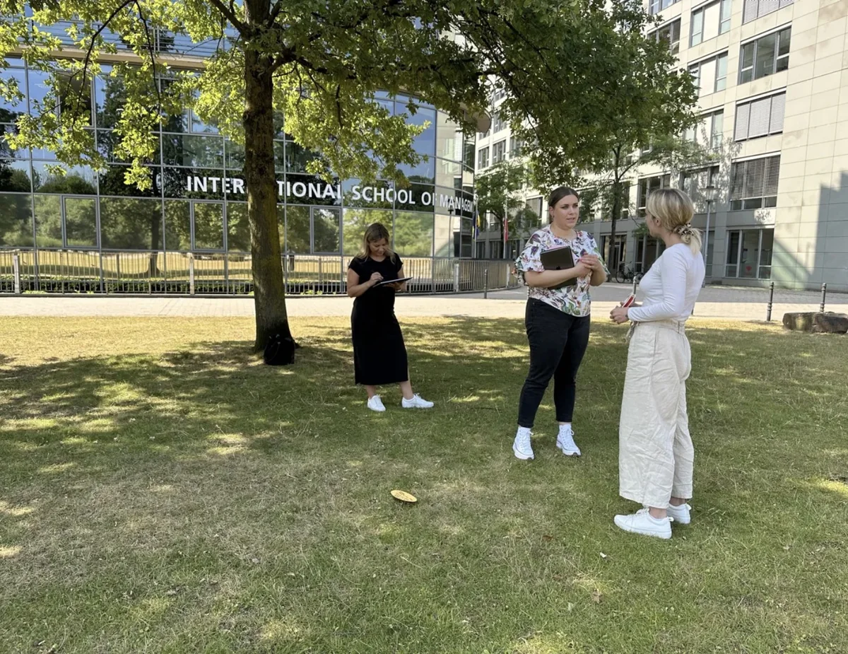 Coaching-Übung zu dritt auf der Wiese vor der International School of Management in Köln, eine Teilnehmerin beobachtet und notiert